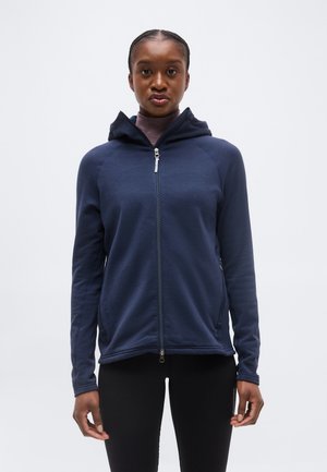 Frau trägt einen marineblauen Kapuzenpullover mit durchgehendem Reißverschluss und Kapuze, schwarze Leggings, steht vor einem schlichten hellen Hintergrund.