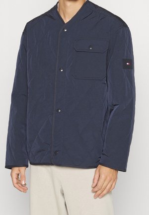 Homme portant une veste matelassée bleu marine avec des boutons sur le devant et une poche poitrine, associée à un pantalon beige clair.