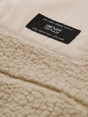 Beige strukturierter Stoff mit einer weichen, flauschigen Oberfläche; besitzt ein schwarzes Etikett mit weißem Text "DENIM TOM TAILOR" und dazugehörigen Details.