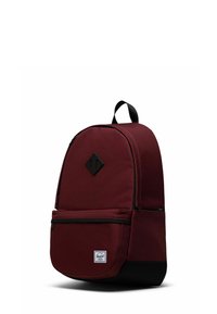 Sac à dos en tissu rouge de forme arrondie, accents noirs, poche avant et écusson logo en forme de losange. Équipé de bretelles réglables.