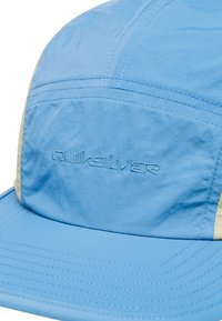 Gorra azul clara hecha de tela texturizada, con acentos crema y costuras visibles. Logotipo bordado de "Quiksilver" en la parte frontal.
