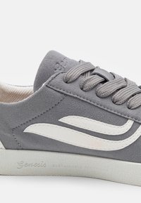 Sapatilha cinza em camurça com detalhes brancos e sola de borracha texturizada. Possui bico arredondado, fecho com atacadores e a marca "Genesis Sustainable Footwear".