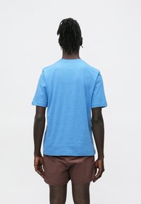 Sinine puuvillast crew-kaelusega T-särk; lühikesed varrukad; tagantvaade. Kantud koos pruunide lühikeste pükstega. Sile tekstuur, minimaalne disain, nähtamatud mustrid.