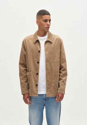 Tan corduroy jacket with a collar, front pockets, and black buttons, worn over a white t-shirt, paired with light blue jeans.