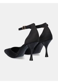 Zapatos de tacón alto de satén negro con puntas afiladas, delgadas correas en el tobillo y tacones en punta elevados, que cuentan con un acabado mate suave y un diseño elegante.