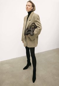 Bēša pārmēru žakete virs melna turtlenecka, apvienota ar melnām legingām un šauriem potīšu zābakiem; rokā tur mazu melnu rokassomu.