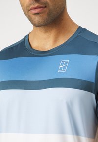 Blå sportskjorte med varierende horisontale striper i mørkeblå, lyseblå og hvit. Har rund hals og en liten logo i øverste venstre hjørne.