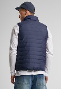 Dunkelblaues gepolstertes Westchen mit hohem Kragen, ausgestattet mit horizontalen Nähten und einer lockeren Passform. Über einem weißen Langarmshirt getragen.