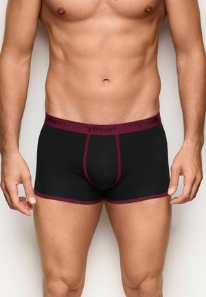 Torsos y muslos masculinos con calzoncillos bóxer negros con cinturilla y ribete granate, de pie sobre un fondo claro y liso.