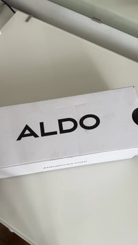 Boîte à chaussures blanche avec la marque "ALDO" en lettres noires en gras sur le dessus. Présente un trou rond pour une ouverture facile et le lien du site web sur le côté.