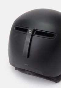 Matně černá helma s hladkým plastovým povrchem, předními ventilačními otvory a vertikálním popruhem. Měkké černé polstrování na spodní straně pro komfort.