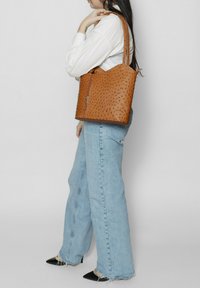 Borsa a tracolla in pelle marrone con motivo testurizzato, chiusura con zip e manici doppi. Abbbinata a jeans a gamba larga blu chiaro e scarpe nere con punta. 