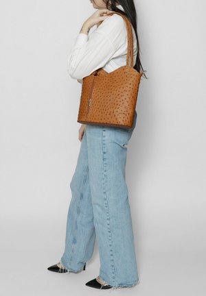 Sac fourre-tout en cuir marron avec un motif texturé, fermeture zippée et poignées doubles. Associé à un pantalon en jean large bleu clair et des chaussures noires à bout pointu.