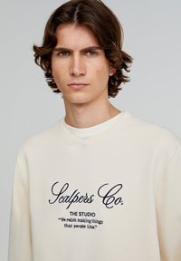 Lichtcrème sweatshirt gemaakt van zachte stof, met geborduurde tekst in donkerblauw. Ronde hals en een relaxte pasvorm zonder patronen.