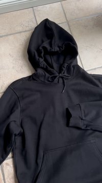 Schwarz-gestreifte Kapuzenpullover mit vorderer Tasche und Kordelzug auf gefliestem Boden, die Ärmel zur Seite abgespreizt.