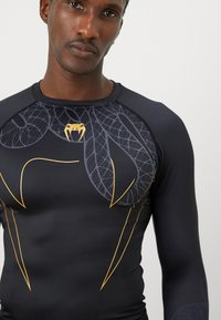 Haut de sport noir à manches longues avec un motif texturé et des accents dorés, offrant une coupe ajustée. Le design comprend un graphique unique à l'avant.
