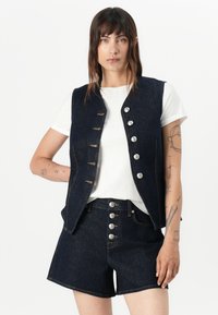 Donkerdenim mouwloos vest met zilveren knopen, gecombineerd met hoge taille donkerdenim shorts. Witte t-shirt eronder, minimalistisch ontwerp.