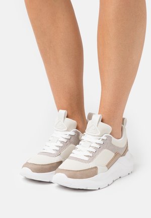 Hvide og beige mesh-sneakers med tykke hvide såler og snørebånd, båret på bare ben mod en ensfarvet baggrund.