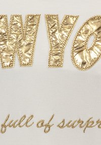 Las letras metálicas doradas dicen "WYOMING" y "lleno de sorpresas" sobre un fondo de tela blanca, con una textura suave y bordados decorativos.