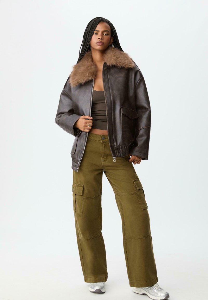 Giacca bomber in pelle marrone con colletto in pelliccia sintetica, abbinata a pantaloni cargo verde oliva con numerose tasche. Modello che indossa sneakers argento.