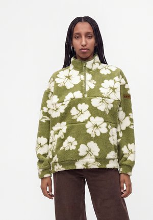Femme aux longs cheveux tressés portant un pull en polaire vert avec un grand motif floral blanc et un pantalon en velours côtelé marron.