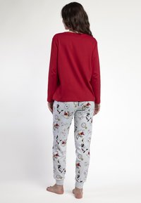 Mujer de pie descalza, de espaldas, vistiendo una camiseta de manga larga roja y pantalones de pijama gris claro con un patrón de Alicia en el país de las maravillas.