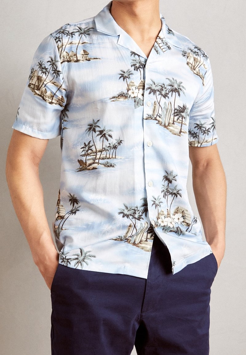 Chemise à manches courtes avec boutons en tissu bleu clair, ornée d'un imprimé tropical avec des palmiers et des planches de surf. Texture lisse, coupe décontractée.