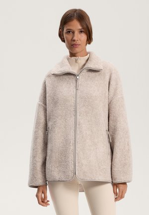 Vloerige beige jas met een hoge kraag en een voorrits. Heeft twee zijzakken en een ontspannen pasvorm. Zachte textuur en casual ontwerp.