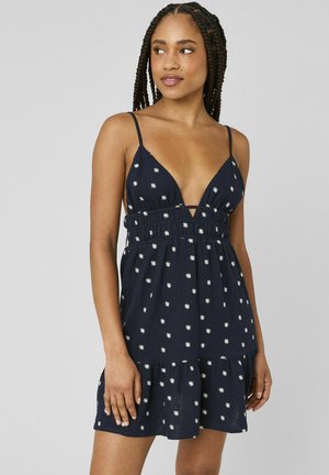 Femme aux cheveux tressés portant une robe sans manches bleu marine à motifs floraux blancs et un décolleté en V profond, debout devant un fond gris.