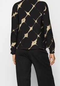 Blusa nera a maniche lunghe con un motivo astratto a diamante beige, caratterizzata da un orlo arrotondato e polsini con bottoni, realizzata in tessuto liscio.