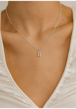 Silver necklace with a delicate chain and a small, textured letter "B" pendant adorned with clear stones, worn on bare skin.