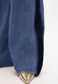 Lepakot denim-housut tummansinisellä sivupaneelilla, joissa on sileä pinta ja kultaiset terävät korkokengät.
