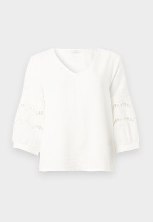 Witte blouse met lange mouwen, V-hals en kanten uitsnijdingen op de mouwen, weergegeven tegen een effen grijze achtergrond.