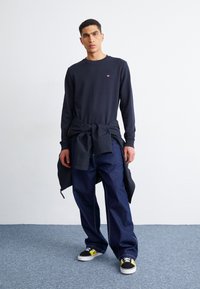 Camicia a maniche lunghe blu marina con un piccolo logo, jeans in denim scuri a vestibilità ampia e sneakers nere con accenti gialli, giacca sovrapposta in vita.