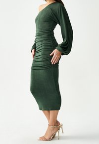 Vihreä, olkapäiltä laskettu mekko, jossa on rypytetty kangas ja pitkät hihat. Keskipitkä siluetti, pehmeä tekstuuri, yhdistettynä kultaisiin korkokenkiin.