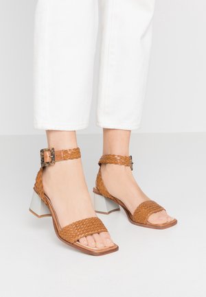 Pieds portant des sandales ouvertes en cuir tressé couleur fauve avec des brides à la cheville et des talons blocs, associées à un pantalon court blanc sur un sol blanc.