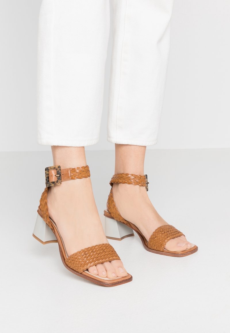 Pieds portant des sandales ouvertes en cuir tressé couleur fauve avec des brides à la cheville et des talons blocs, associées à un pantalon court blanc sur un sol blanc.
