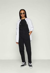 Woman wearing black overalls, black shirt, white open button-up shirt, black and white sneakers, and round sunglasses, standing against plain background.