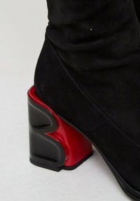 Botín de ante negro con un tacón grueso que presenta un diseño geométrico único en negro y rojo. El tacón tiene un acabado brillante.