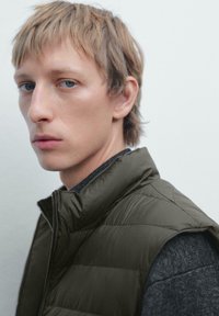 Ung person med kort, lyst hår iført en mørkegrøn polstret vest og en mørk strikket sweater, vender let til venstre mod en ensfarvet baggrund.