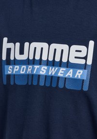 Granatowa tkanina z białym napisem "hummel" i niebieską metką "SPORTSWEAR", z pionowymi niebieskimi paskowanymi wzorami za tekstem.