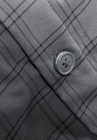 Tessuto a quadri grigio con una sottile texture, caratterizzato da un pulsante grigio scuro impresso con la scritta "Mammut". Vista ravvicinata che mette in evidenza i motivi di tessitura.