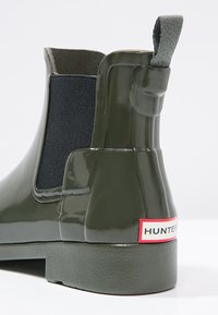 Botte de pluie brillante vert foncé à la cheville avec panneau latéral élastique noir et languette, avec étiquette "Hunter" sur le talon.