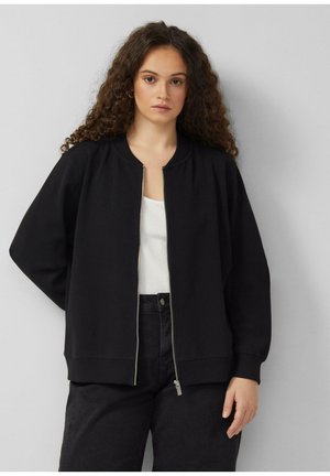 Frau mit lockigem Haar, die eine schwarze Zip-up-Jacke über einem weißen Oberteil und schwarze Hosen trägt, steht vor einem schlichten hellgrauen Hintergrund.