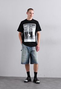 Tricou negru Moschino cu imprimeu grafic, pantaloni scurți din denim decolorat albastru deschis, șosete negre și pantofi negri lucioși.