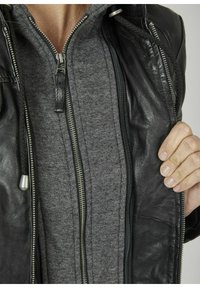 Schwarze Lederjacke mit silbernen Reißverschlüssen, getragen über einem grauen Kapuzenpullover mit Reißverschluss. Nahaufnahme zeigt Texturdetails und geschichtete Stoffe.