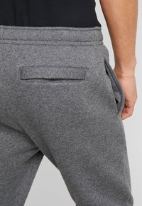 Nahaufnahme einer Person in grauen Sweatpants mit einer Klappe an der Gesäßtasche und einer Hand in der Seitentasche vor einem schlichten Hintergrund.