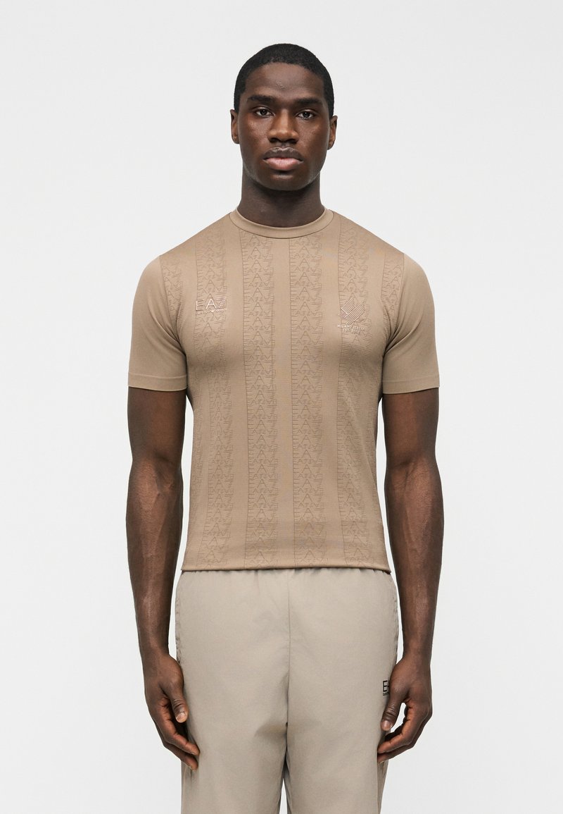Άνδρας που φοράει μπεζ πουκάμισο με κοντά μανίκια και διακριτικά επαναλαμβανόμενα μοτίβα με το λογότυπο EA7, καθώς και ανοιχτό μπεζ παντελόνι, σε απλό λευκό φόντο.