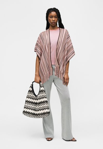 Femme portant un châle à motif zigzag rose, un haut rose clair, un pantalon large gris, des sandales noires, tenant un grand sac à motif zigzag noir et blanc.