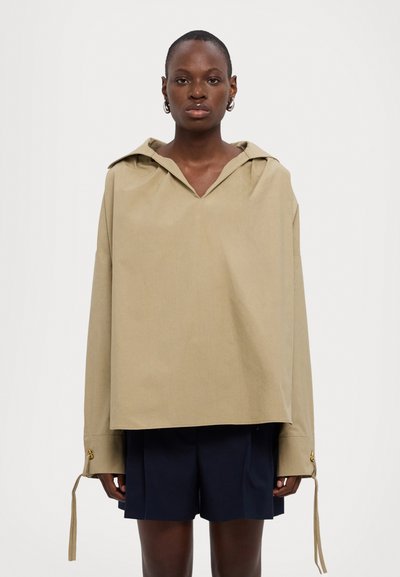 Maglione khaki oversize con cappuccio, scollo a V profondo, maniche lunghe e polsini regolabili con dettagli dorati. Indossato su pantaloni corti blu navy.